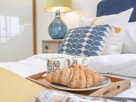 Una cama impecablemente hecha con ropa blanca, una lámpara azul y una bandeja con un croissant y dos tazas de café sobre una bandeja de madera.
