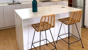 A modern kitchen with a central island, two bar stools, pendant lights, a refrigerator, and a window with natural light. - Villa Tres Mares - Extraordinary accommodation. (Poblado de Sancti Petri)