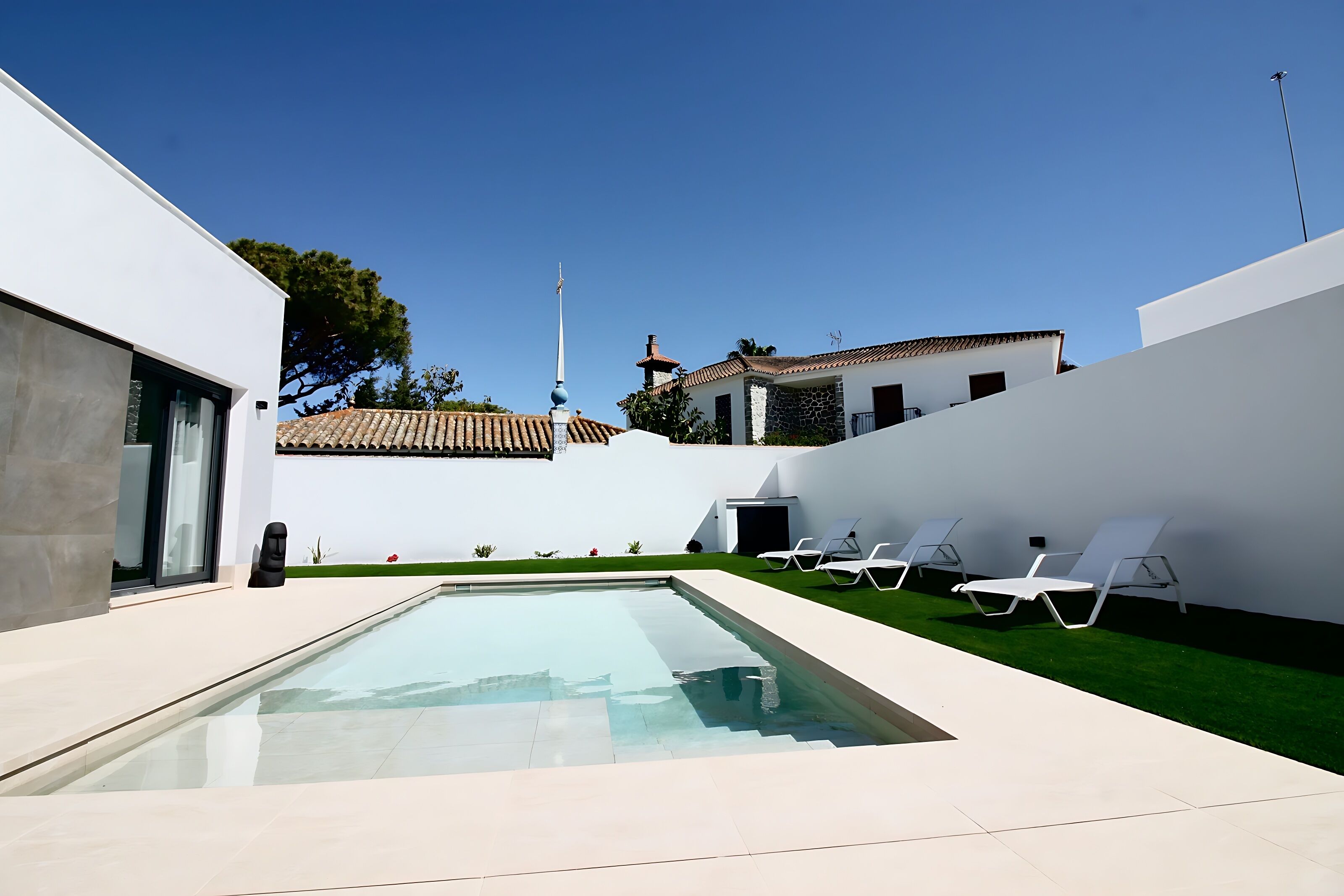 A modern pool area with a clear pool, white lounge chairs, and a view of traditional buildings in the background.