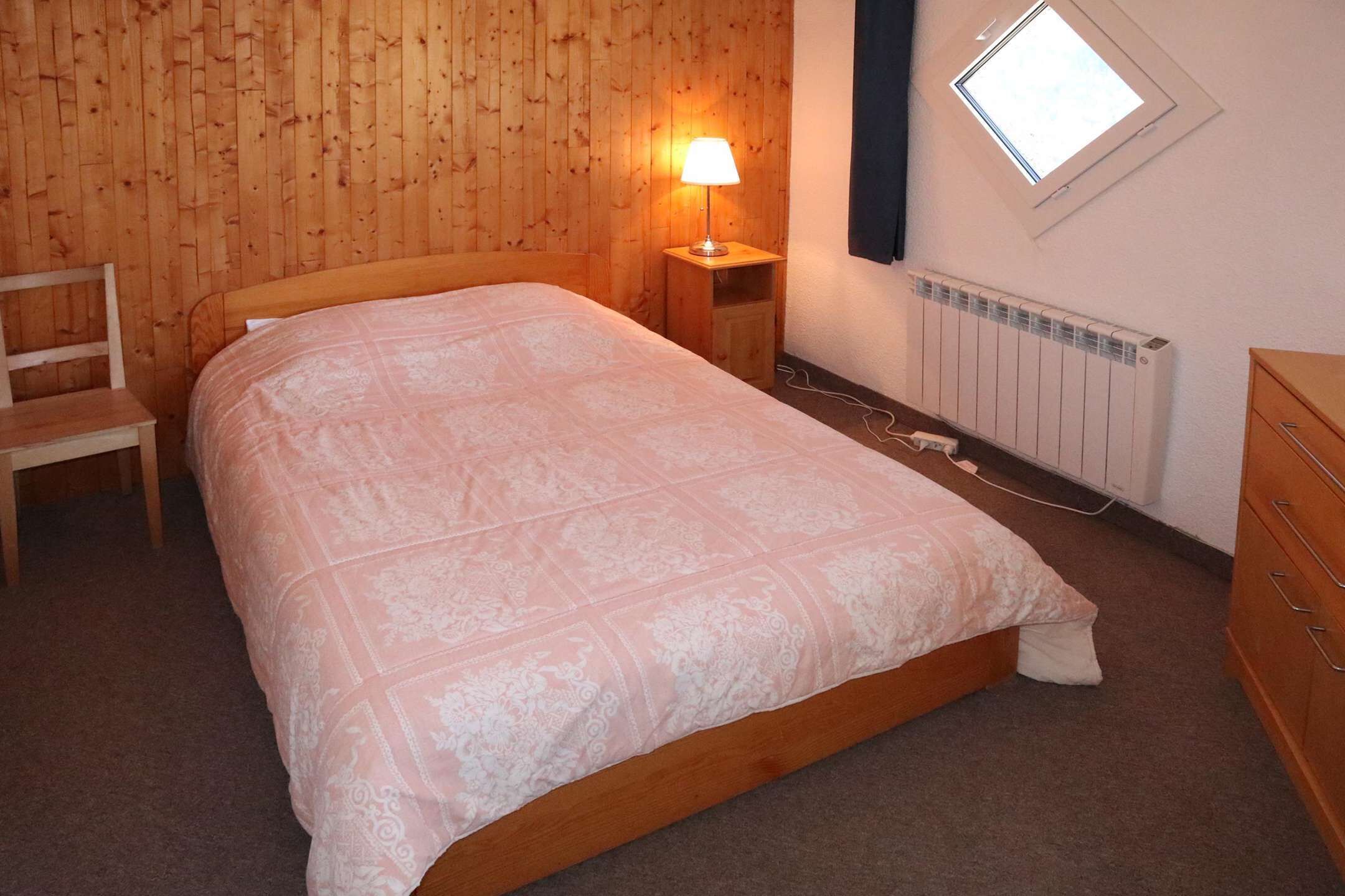 A bedroom with a wooden headboard, a single bed with a patterned bedspread, a bedside table with a lamp, a radiator, and a window with a view.