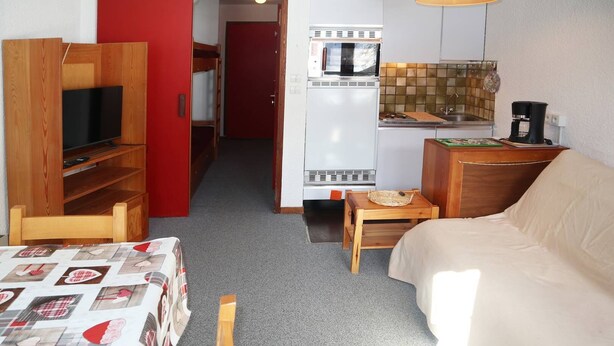 A compact room with a sofa, a table with a patterned tablecloth, a kitchenette with a microwave and coffee maker, and a wooden TV stand. - Départ Skis aux Pieds (LES ORRES 1650)