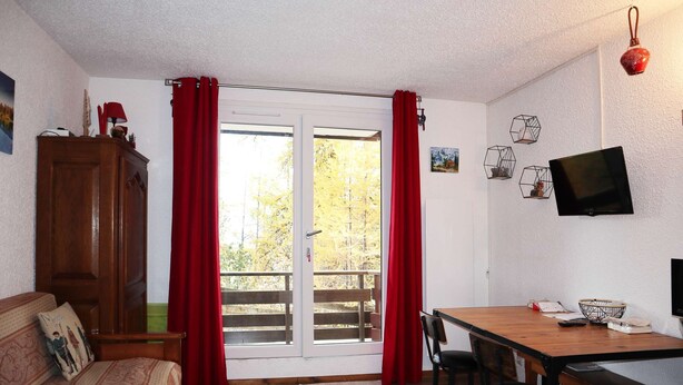 A room with a wooden dining table, two chairs, a sofa with patterned cushions, a wooden cabinet, and a television on the wall. - Studio 6 Personnes Accès Direct aux Pistes (LES ORRES 1650)