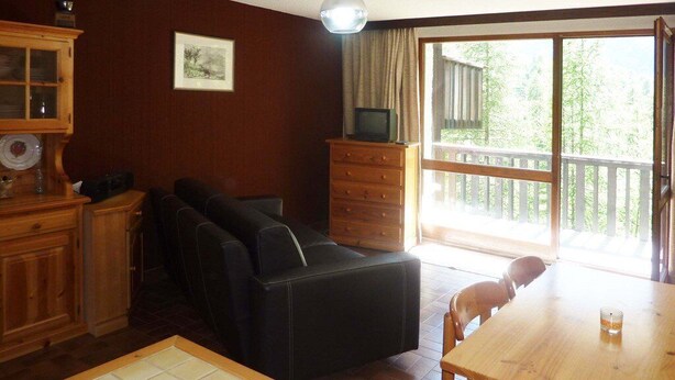Ein Zimmer mit einem Holzespeisetisch, einer schwarzen Couch, einem Holzkabinett und einem Fernseher auf einer Kommode. Ein großes Fenster bietet Blick auf Bäume.