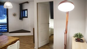 Ein Zimmer mit Holzboden, einem Esstisch mit Stühlen, einer Stehlampe, einem kleinen Tisch mit Pflanze und einer Tür zu einem anderen Raum.