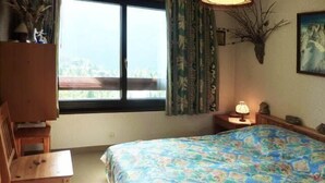 A bedroom with a bed, a desk, a chair, a window with curtains, a lamp, and a painting on the wall.