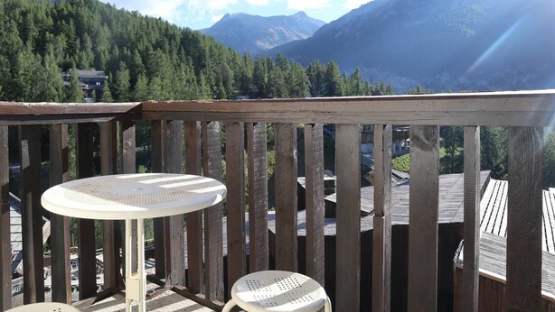 En træterrasse med et rundt bord og to stole, med udsigt over et bjergrigt landskab under en klar himmel.
