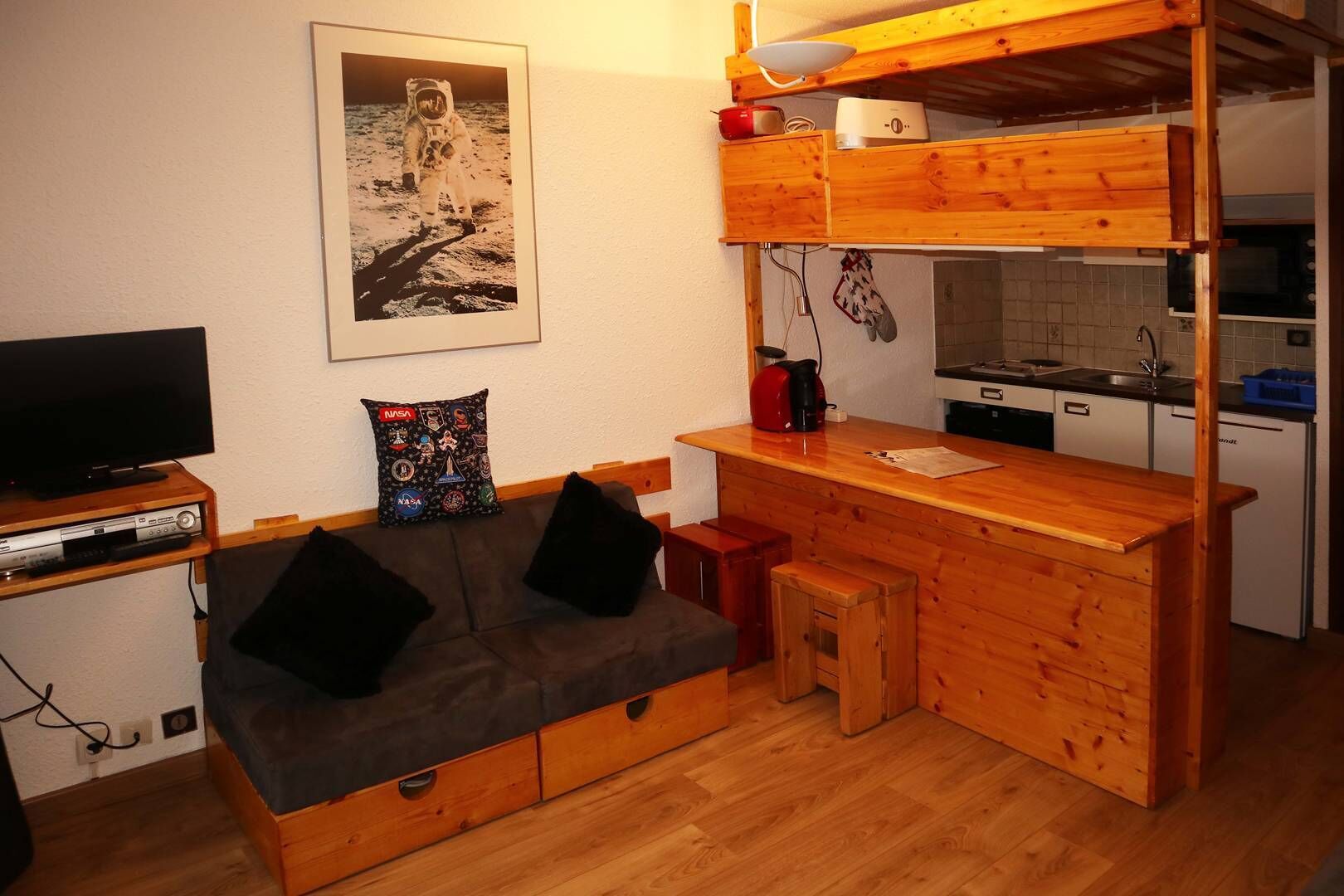 A compact living space with a kitchenette, a sofa with storage drawers, a television, and a NASA-themed pillow.