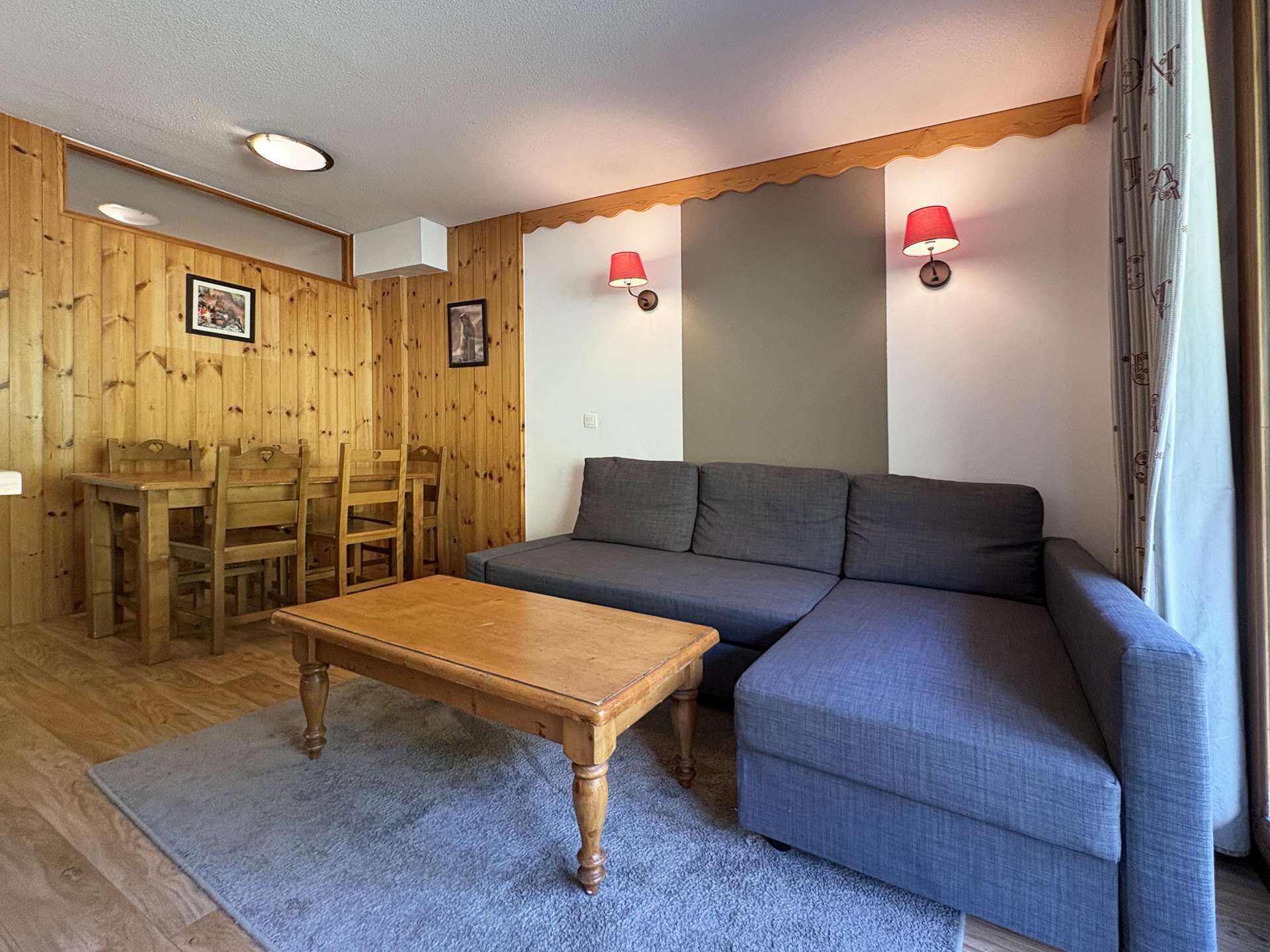 A room with a wooden dining table, chairs, a sofa, and two wall-mounted lamps.