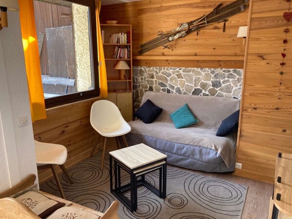 Interior acolhedor de uma cabana, com parede de pedra, móveis de madeira, estante de livros e esquis na parede.
