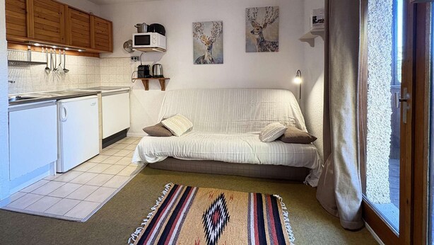 A compact kitchen with white appliances, a sofa with cushions, and artwork on the walls. - Studio à Deux pas du Centre (LES ORRES 1650)