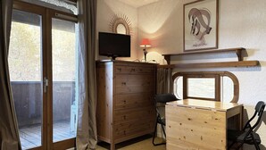 A cozy room with a sliding glass door leading to a balcony, a wooden dresser, a desk with a chair, and a framed artwork on the wall. - Studio à Deux pas du Centre (LES ORRES 1650)