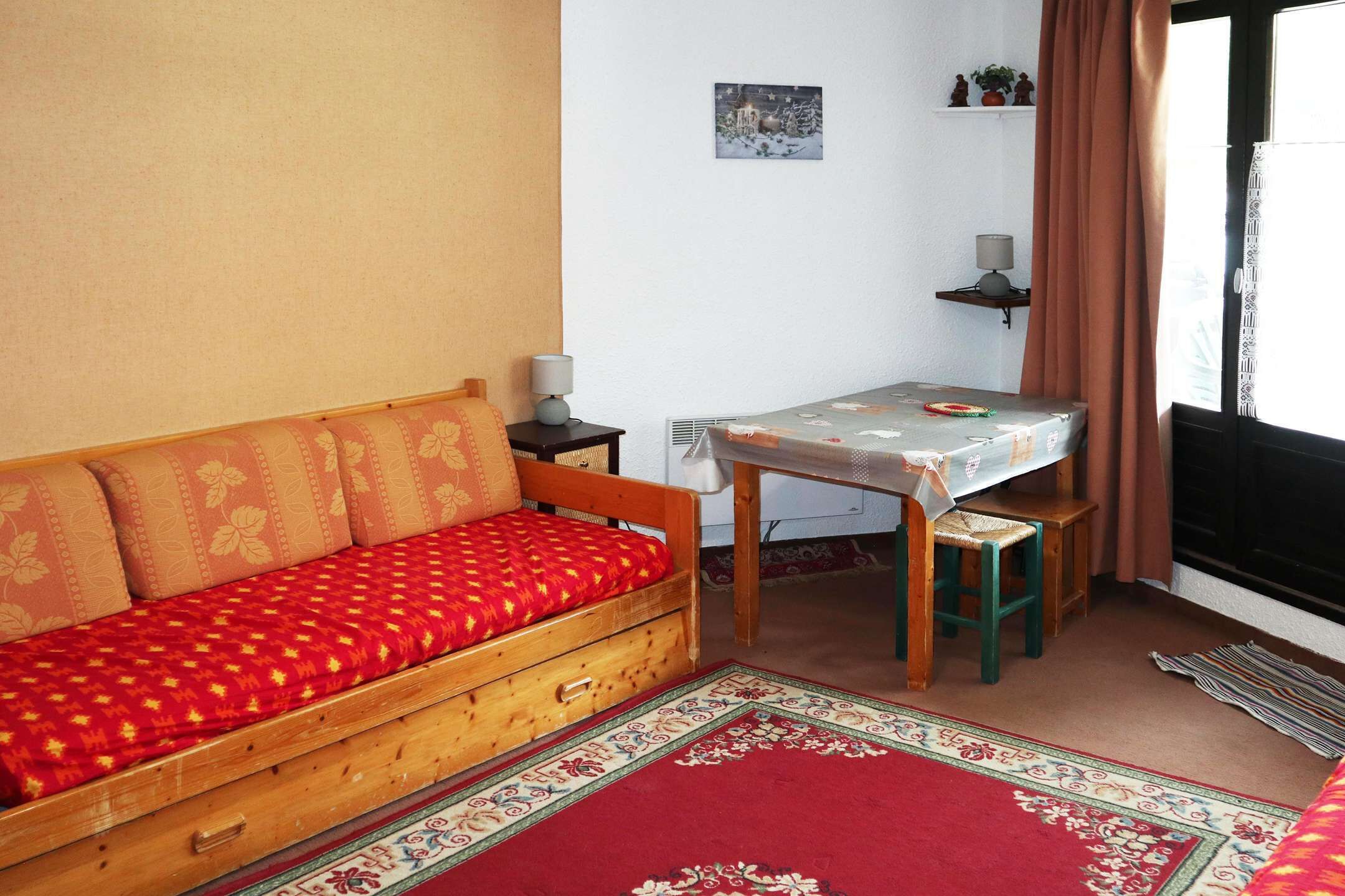 A small room with a wooden sofa, a small table with a tablecloth, a chair, a radiator, a window with curtains, and a wall-mounted shelf with a lamp.