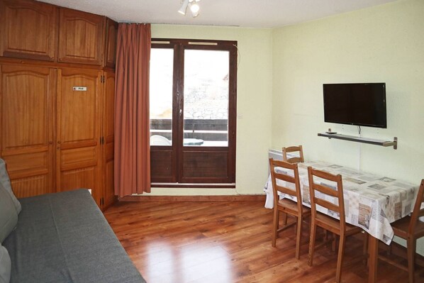 A room with wooden flooring, a sofa, a dining table with chairs, a television mounted on the wall, and a sliding glass door leading to an outdoor area. - Studio Équipé Pour 4 Personnes vue Pistes (LES ORRES 1650)