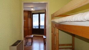 Une chambre avec un lit superposé, une petite table et un banc. Une porte donne sur un balcon avec vue.