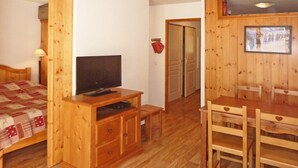 Una habitación con piso de madera, una cama, un televisor sobre un atril de madera, una mesa de comedor con sillas y una pared de madera con un cuadro enmarcado.