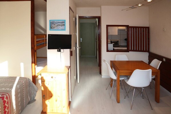 A compact room with a bed, a desk with a flat-screen TV, a dining table with chairs, a kitchenette, and a mirror.