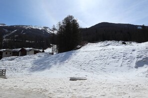 A snow-covered landscape with a wooden bench, a small building, and a mountain in the background.