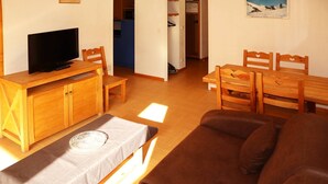 A cozy living room with a brown sofa, wooden TV stand, and a dining table with chairs. - Appartement Parc DES Airelles 211 (LES ORRES 1800)