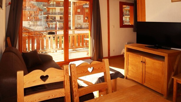 A cozy living room with a sofa, wooden chairs, a TV on a wooden cabinet, and a large sliding glass door leading to a balcony with wooden railings. - Appartement Parc DES Airelles 211 (LES ORRES 1800)
