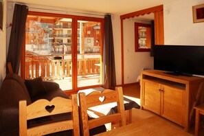 A cozy living room with a sofa, wooden chairs, a TV on a wooden cabinet, and a large sliding glass door leading to a balcony with wooden railings. - Appartement Parc DES Airelles 211 (LES ORRES 1800)