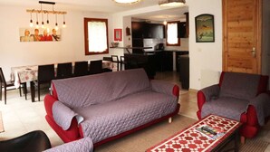 Uma sala de estar com sofá, poltronas, mesa de centro e um espaço de jantar com cadeiras e mesa. Cozinha visível ao fundo.