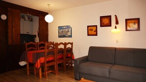 Ein Wohnzimmer mit grauem Sofa, Esstisch aus Holz mit Stühlen, einem Holztisch und einer Wanduhr.