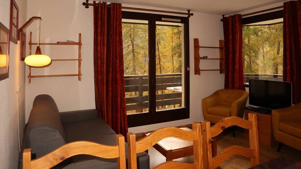 Ein gemütliches Zimmer mit einer Couch, Holzstühlen, einem Fernseher und großen Fenstern mit Vorhängen.