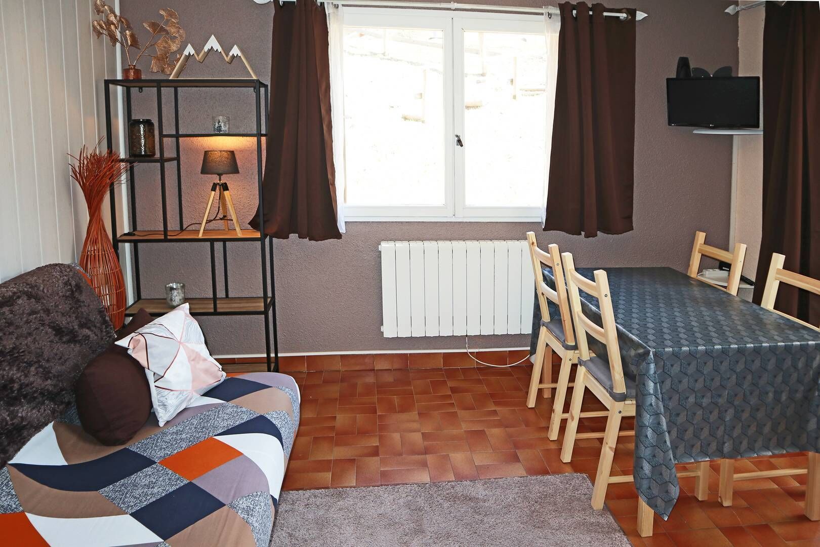 A cozy living room with a sofa, a dining table with chairs, a TV mounted on the wall, and a window with curtains.