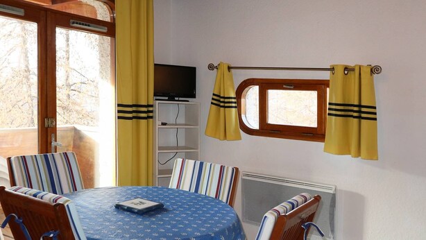 A dining area with a round table, striped chairs, a TV on a shelf, and yellow curtains. - Studio Pour 4 Personnes Rénové Avec Soin (LES ORRES 1650)