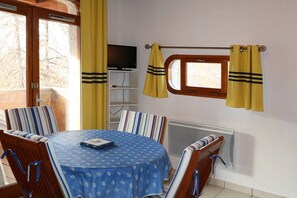 A dining area with a round table, striped chairs, a TV on a shelf, and yellow curtains. - Studio Pour 4 Personnes Rénové Avec Soin (LES ORRES 1650)