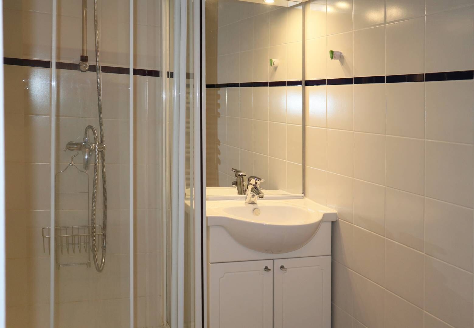Ein Badezimmer mit einer gläsernen Duschkabine, einem weißen Waschbecken inklusive Spiegel und einer gefliesten Wand mit einem schwarzen Streifen.
