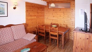 L’intérieur d’un chalet accueillant, avec un mur en bois, un canapé, une table à manger entourée de chaises, une petite table sur laquelle se trouve un bol, un téléviseur posé sur un meuble et une lampe.