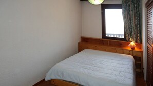 A small, single-bed room with a wooden headboard, a bedside lamp, and a window with curtains. - Appartement G307 LES Cembros (LES ORRES 1650)