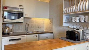 A compact kitchen with white cabinets, a Brandt microwave, a coffee maker, a toaster, and a dish rack with white dishes.
