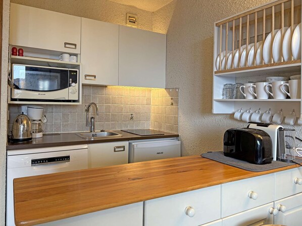 Una cocina compacta con gabinetes blancos, un microondas Brandt, cafetera, tostadora y un escurreplatos con vajilla blanca.