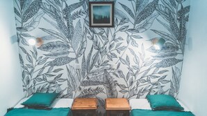 Een kamer met behangpapier met bladmotieven, twee houten nachtkastjes, slaapgedeelte in teal-kleur en een ingelijst kunstwerk aan de muur.