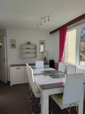 Een eethoek met een tafel en stoelen, een klein plankje met decoratieve objecten, een klok en een raam met gordijnen.
