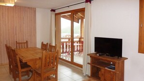 Una mesa de comedor de madera con sillas, un televisor sobre un mueble de madera y un gran ventanal corredizo que da a un espacio exterior con sillas rojas.