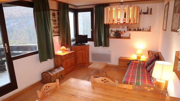 A cozy room with a wooden dining table, a small wooden cabinet, a television on a wooden stand, a sofa with a plaid blanket, and a shelf with books and decorative items.