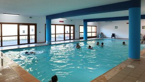 An indoor swimming pool with clear glass walls, blue pillars, and tiled flooring.
