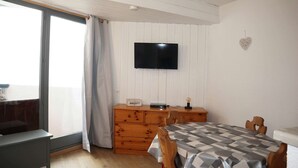 A room with a wooden dresser, a television mounted on the wall, a table with a checkered tablecloth, and a window with curtains.