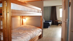 A bunk bed with a headboard and a bedside table, a sofa, a small table, and a cabinet in a room with wooden flooring and a door leading to another room. - Studio 4 Personnes en Cœur de Station (LES ORRES 1650)