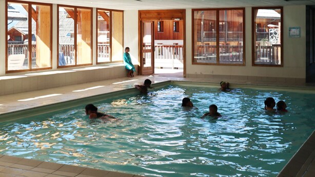 An indoor swimming pool with clear views through large windows, wooden deck, and a person sitting by the pool.