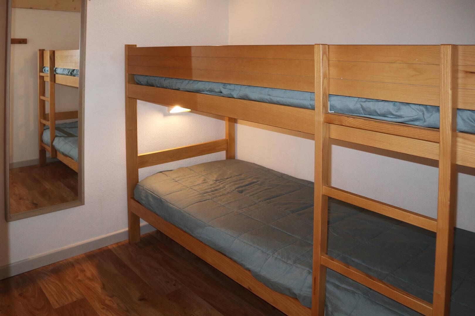 A bunk bed with a mirror on the left side, wooden flooring, and a light source on the left.