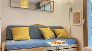 A room with a blue sofa, three yellow cushions, a small table with a tray of fruit and two glasses of beer, a mirror, and a lamp.