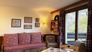 A cozy room with a checkered sofa, a wooden table with a heart-patterned tablecloth, and a window with a view of trees.