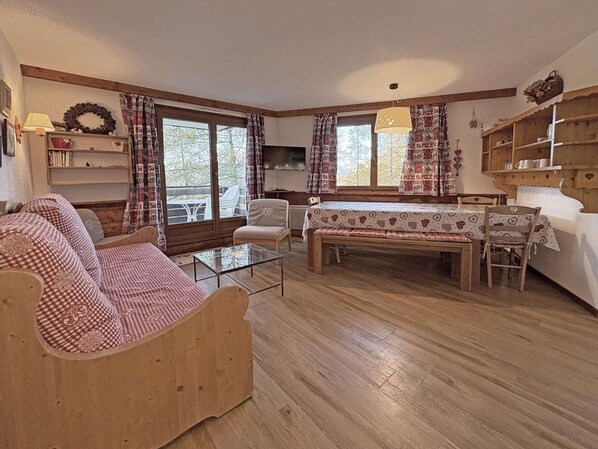 A cozy living room with a wooden sofa, a glass coffee table, and a dining table with chairs. There are curtains, a fireplace, and a bookshelf. - BEL APPARTEMENT 4 PIECES POUR 10 PERSONNES (LES ORRES 1650)