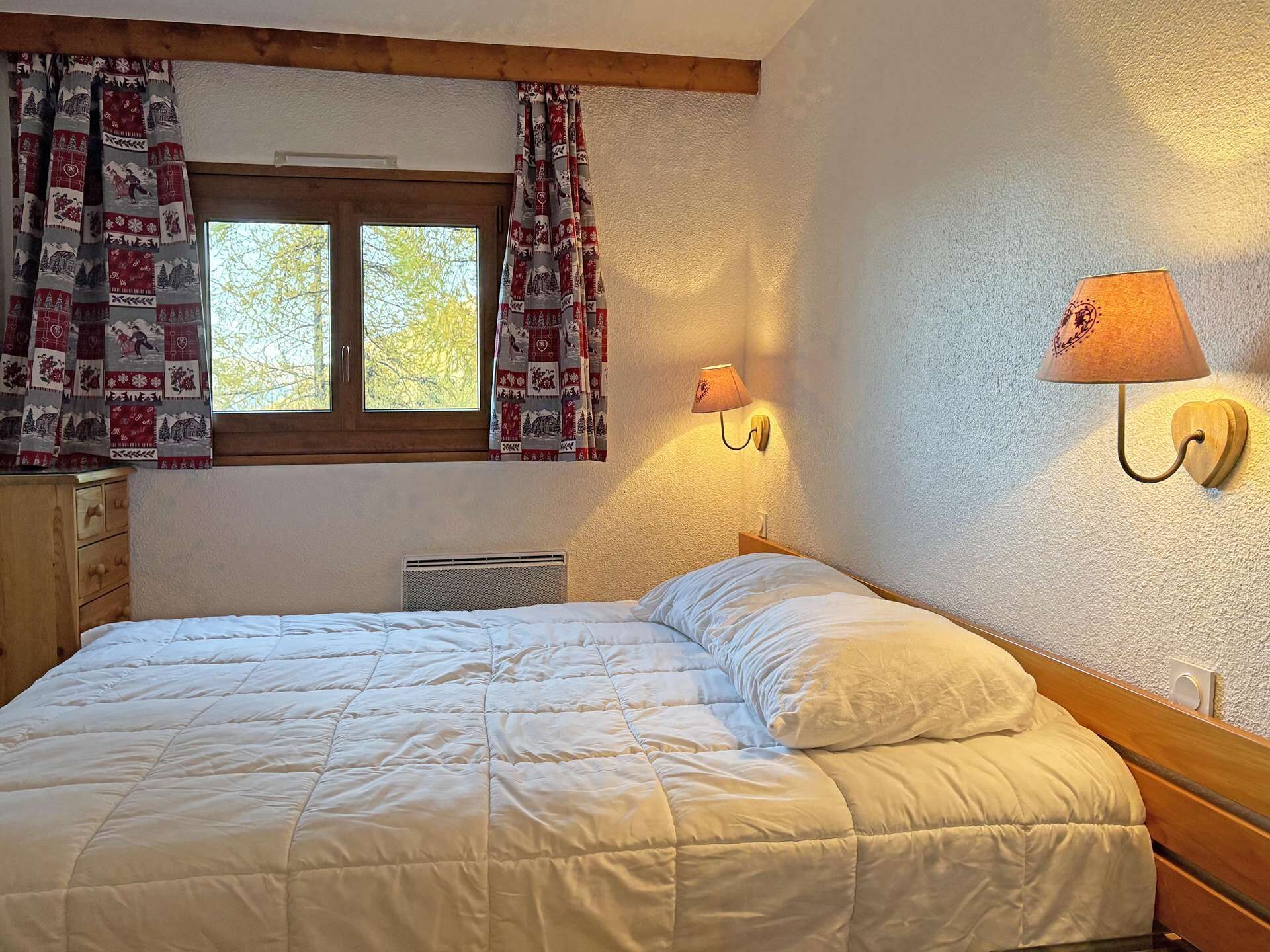 A bedroom with a bed, a window with curtains, a wall-mounted lamp, and a wooden dresser.