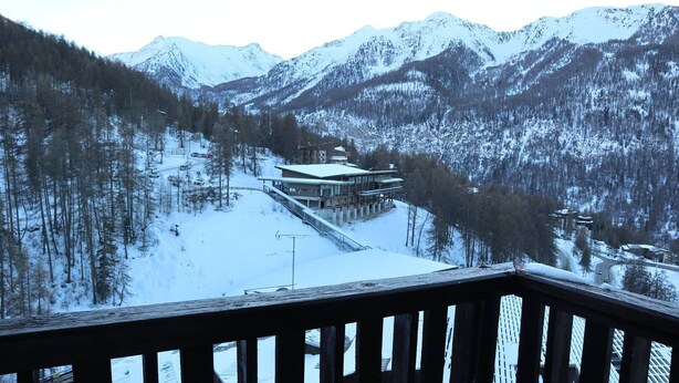 Un paysage montagneux enneigé, avec un bâtiment doté d’une façade en verre et d’une terrasse en bois qui s’étend sur une pente couverte de neige.