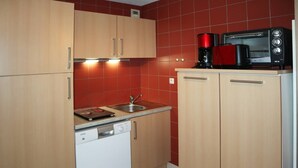 A compact kitchen with wooden cabinets, a white dishwasher, a red-tiled backsplash, and a coffee maker on the countertop. - Studio N° 211 LES Flocons (LES ORRES 1650)